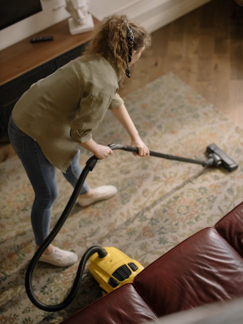 Close-up image of a black vacuum cleaner with a slim nozzle attachment, actively cleaning a traditional patterned area rug with floral motifs in soft hues of beige, green, and orange. The rug is laid