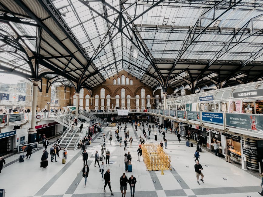 Interior of a busy train station with a high glass and steel arched ceiling, large windows, and natural light illuminating the spacious main hall. The polished tiled floor appears clean and well-maint
