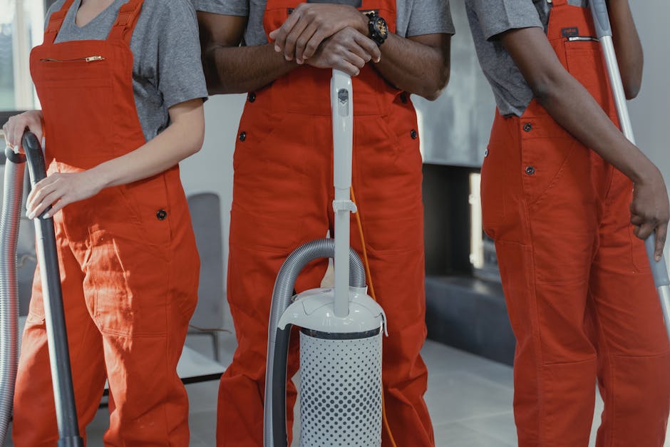 Three professional cleaners from Somers Town Carpet Cleaners, dressed in gray shirts and orange overalls, are holding vacuum cleaners and cleaning tools in a room with wooden floors, ceramic tiles, an