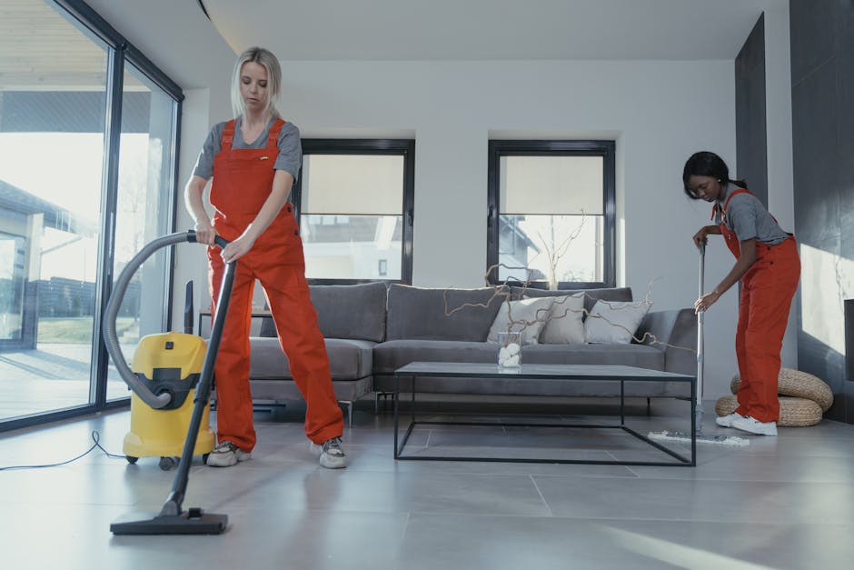 Three professional cleaners from Somers Town Carpet Cleaners, dressed in gray shirts and orange overalls, are holding vacuum cleaners and cleaning tools in a room with wooden floors, ceramic tiles, an